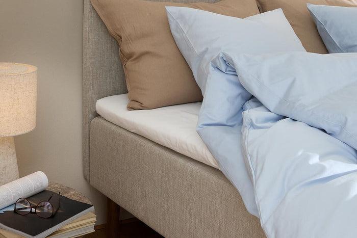 Beige seng med lyseblåt sengetøj og brune puder ved siden af et sengebord med lampe, bøger og briller i blødt dagslys.
