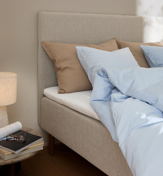 Beige seng med lyseblåt sengetøj og brune puder ved siden af et sengebord med lampe, bøger og briller i blødt dagslys.