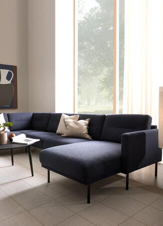 Moderne stue med mørkeblå hjørnesofa med chaiselong, sort sofabord med plante, væghylder og abstrakt vasebillede ved stort vindue med lyse gardiner i blødt dagslys.