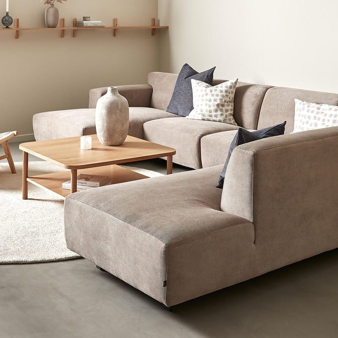 Beige hjørnesofa med træ-sofabord, neutrale puder og væghylde med vase og lys i en lys stue.