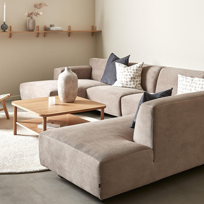 Beige hjørnesofa med træ-sofabord, neutrale puder og væghylde med vase og lys i en lys stue.