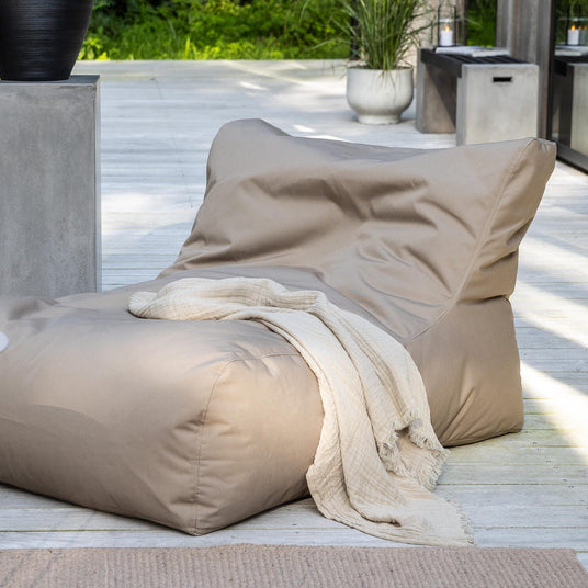 Beige lounge-sækkestol på træterrasse med lys plaid, rundt sidebord med lysestage og krukker med grønne planter i dagslys.