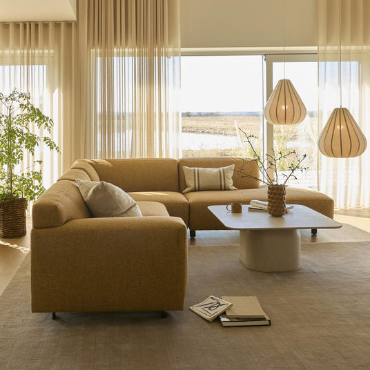 Lyst stue med beige hjørnesofa, lavt sofabord med vase og kop, to pendler og lette gardiner i dagslys.