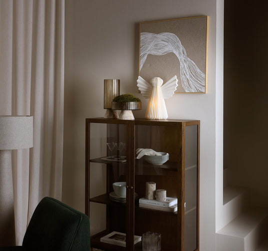 Vitrineskab i mørkt træ med glaslåger, engelformet lampe og vase med mos ovenpå, abstrakt vægbillede, bogen “Kinfolk” på hylden samt skåle, glas og lys; beige gardiner og varm, dæmpet belysning.
