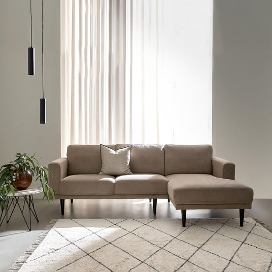 Beige hjørnesofa med chaiselong i en lys, minimalistisk stue med lette gardiner, to sorte pendler, et lille sidebord med grøn plante og et lyst tæppe med rudemønster.