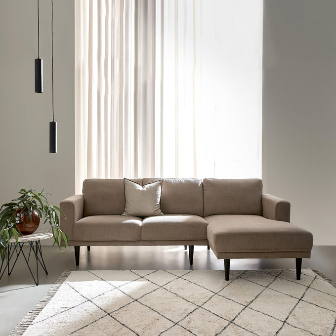 Beige hjørnesofa med chaiselong i en lys, minimalistisk stue med lette gardiner, to sorte pendler, et lille sidebord med grøn plante og et lyst tæppe med rudemønster.
