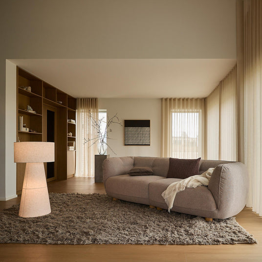 Lys stue med beige gardiner, stor brunbeige sofa på blødt ryatæppe, gulvlampe og indbyggede reoler ved store vinduer i dagslys.