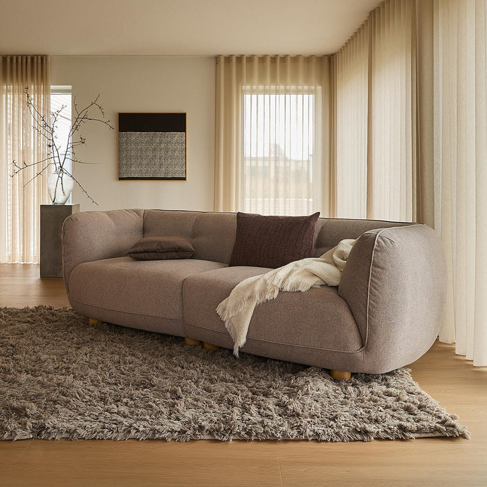 Lys stue med beige gardiner, stor brunbeige sofa på blødt ryatæppe, gulvlampe og indbyggede reoler ved store vinduer i dagslys.