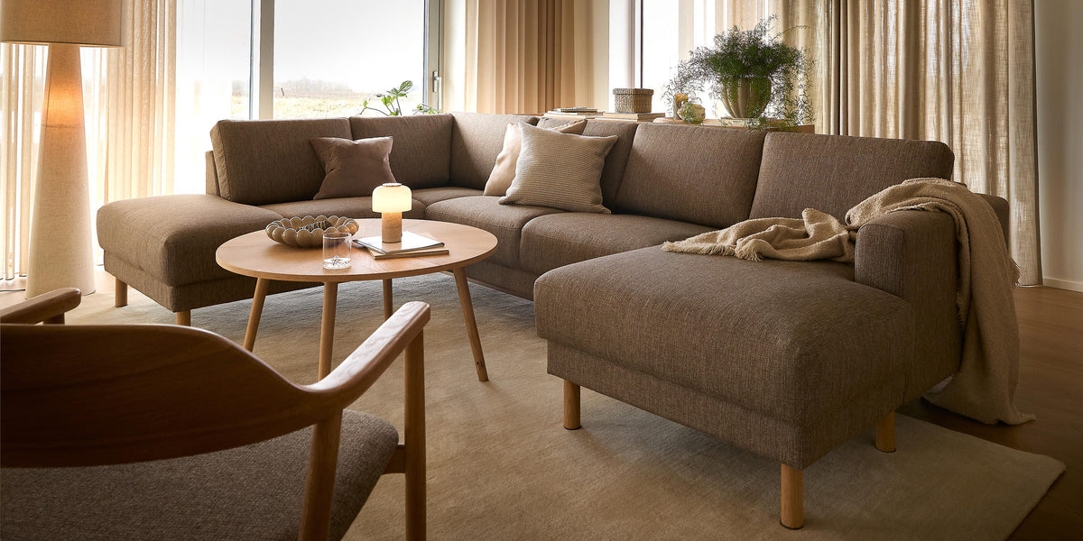 Lys stue med beige gardiner, brun hjørnesofa og rundt sofabord i træ med tænd bordlampe og bog.