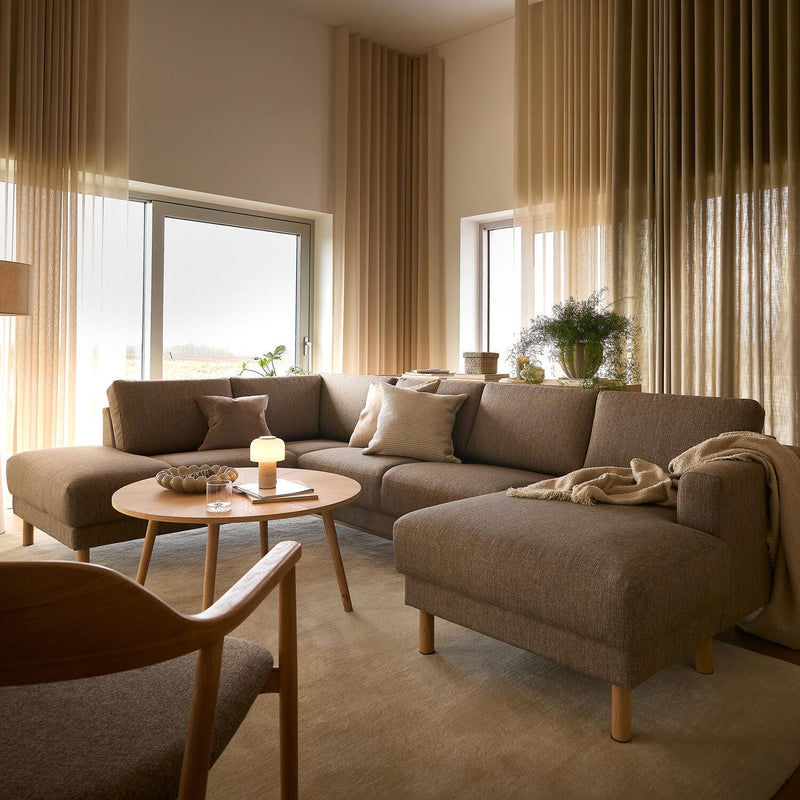 Lys stue med beige gardiner, brun hjørnesofa og rundt sofabord i træ med tænd bordlampe og bog.