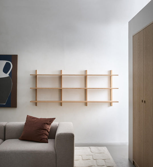 Minimalistisk stue med væghylde i lyst egetræ over en grå sofa, brun pude og lyst tæppe.