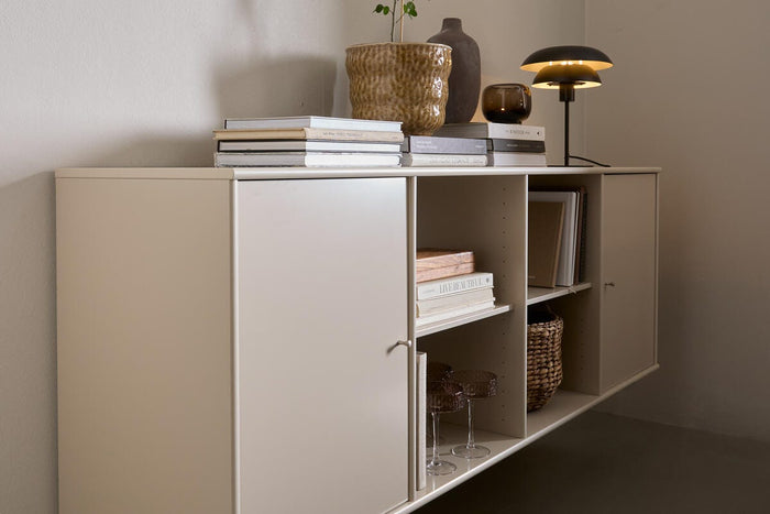 Beige vægmonteret skab med åbne hylder, bøger, bordlampe og krukke med grøn gren.