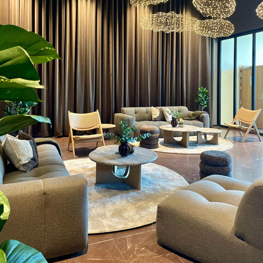 Moderne lounge med taupe modulsofaer, runde tæpper, træ-sofaborde og skyformede pendler foran høje brune gardiner og grønne planter.