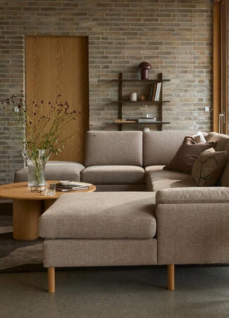 Stue med L-formet beige sofa, træborde, gulvlampe og glasvase med grene; store vinduer og murstensvæg.