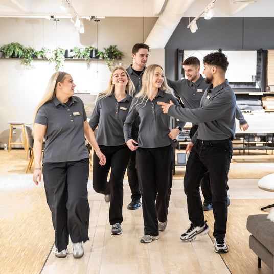 Gruppe butikmedarbejdere i mørkegrå uniform går sammen gennem et møbelshowroom.