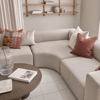 Beige modulsofa med buet hjørne og rust- samt hvide puder, rundt sofabord i mørkt træ med bog og glasvase, åbne hylder med keramik og bøger, lyse gardiner og tæppe i en skandinavisk stue.
