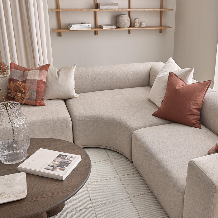 Beige modulsofa med buet hjørne og rust- samt hvide puder, rundt sofabord i mørkt træ med bog og glasvase, åbne hylder med keramik og bøger, lyse gardiner og tæppe i en skandinavisk stue.