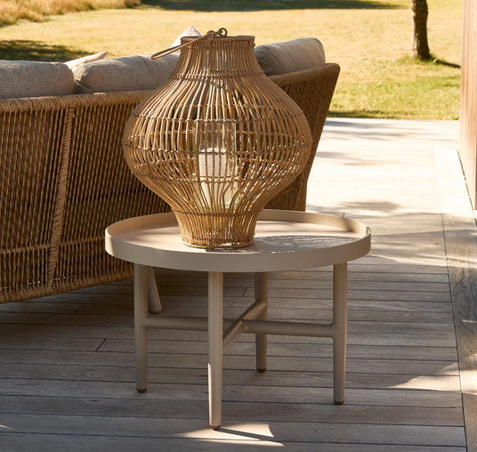Rundt beige udendørs sidebord med en stor fletlanterne ovenpå ved siden af en fletsofa på en træterrasse i sollys med græs i baggrunden.