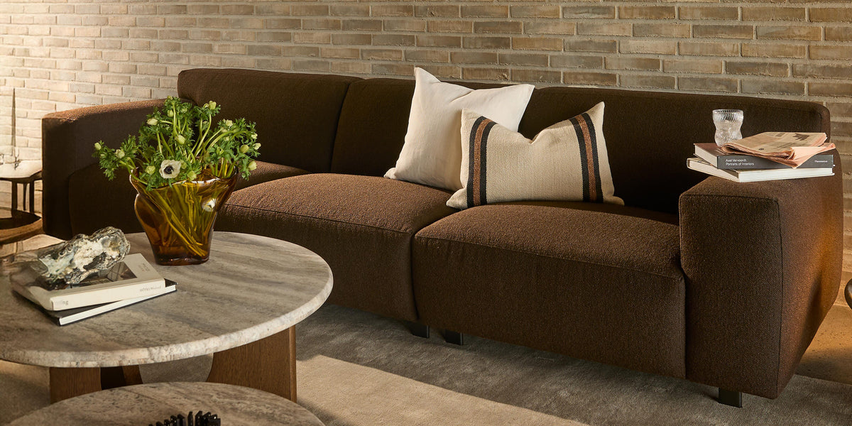 Brun sofa foran lys murstensvæg i stue med to runde stentop-borde, glasvase med grønne blomster, puder, bøger og et glas.
