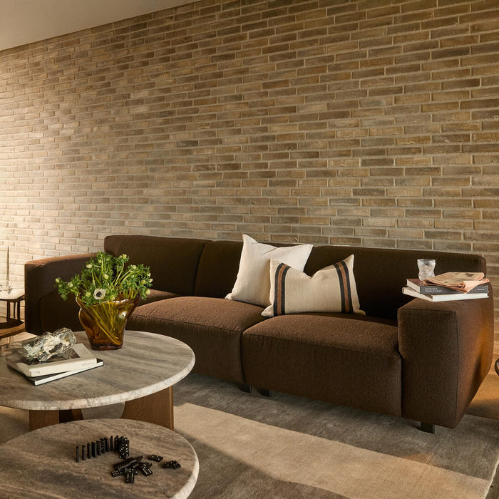 Brun sofa foran lys murstensvæg i stue med to runde stentop-borde, glasvase med grønne blomster, puder, bøger og et glas.