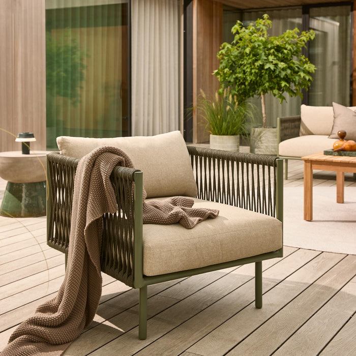 Udendørs lænestol med beige hynder og en strikket plaid på en træterrasse, omgivet af potteplanter og et lavt bord i en moderne patio.