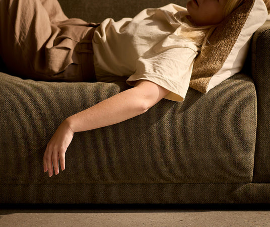 Person der hviler på en olivengrøn sofa med armen hængende ned over kanten, varmt sollys og beige pude.