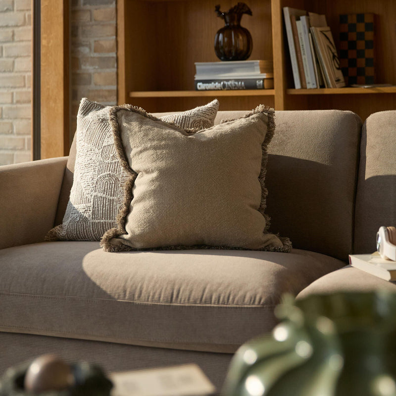 Beige sofa med to puder med frynser i varmt sollys, træreol med bøger og vase i baggrunden.