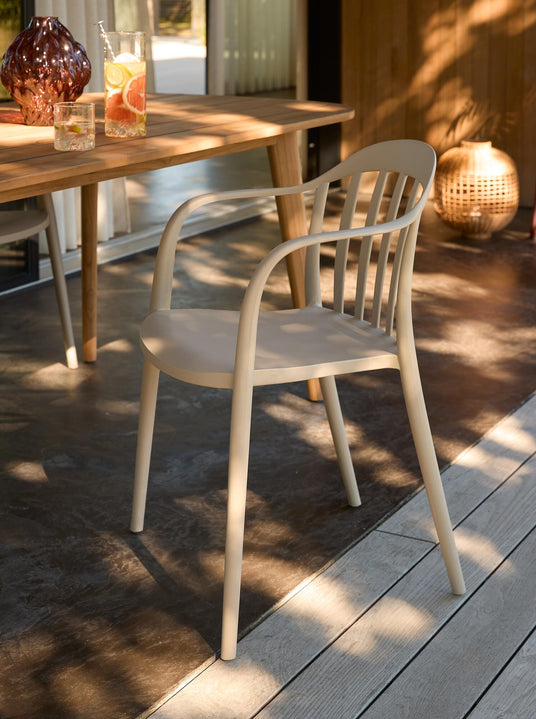 Beige udendørs stol med armlæn ved et træbord på terrassen, solpletter på gulvet og en kande med citrondrik og glas på bordet.