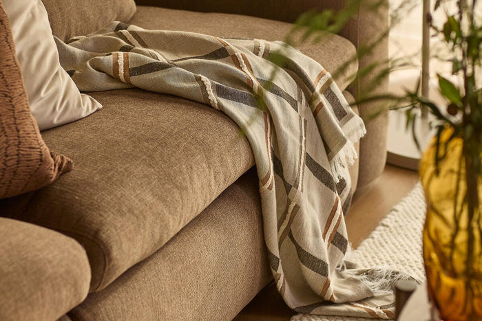 Nærbillede af beige/brun sofa med stribet plaid og puder; ravfarvet vase og lyst, struktureret tæppe i en varm stue.