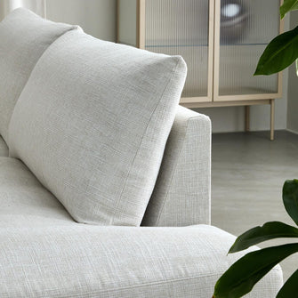 Nærbillede af lys beige stofsofa med grov væv, grøn plante i forgrunden og et lyst skab i baggrunden.