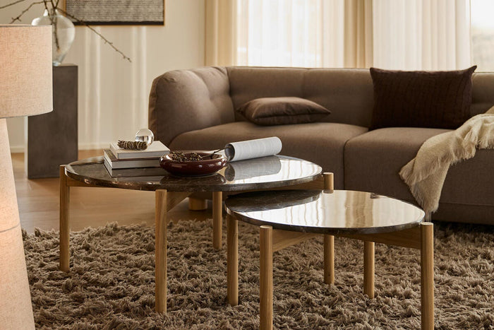 Hyggeligt stue-miljø med to ovale sæt-borde med mørke stenplader og træben på et langhåret tæppe foran en beige sofa i blødt dagslys.