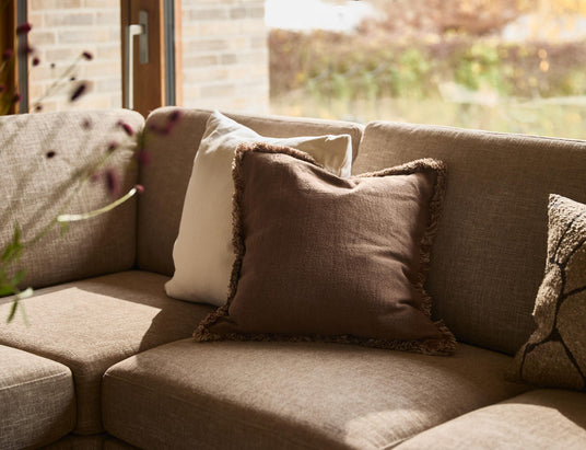 Nærbillede af en beige stofsofa med en brun frynset pude og lyse puder ved et stort vindue i varmt dagslys, med sløret udsyn til haven.