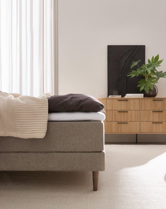 Lyst soveværelse med beige polstret seng med strikket plaid og mørk pude, trækommode med grøn plante og sort kunstværk i blødt dagslys fra et vindue med gardiner.