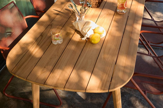 Udendørs teakbord med røde netstole, citroner og kande med citrusvand i sollys.