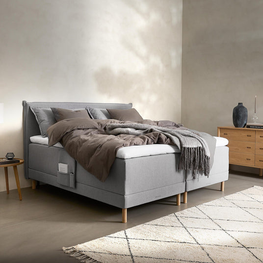Lyst soveværelse med grå kontinentalseng på træben, sengetøj i beige og grå nuancer med plaid over kanten, lyst ternet tæppe og trækommode med vase.