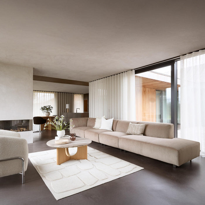 Lys, minimalistisk stue med beige modulsofa, hvid struktureret tæppe, træ-sofabord med vase og blomster, store skydedøre i glas og lette gardiner.