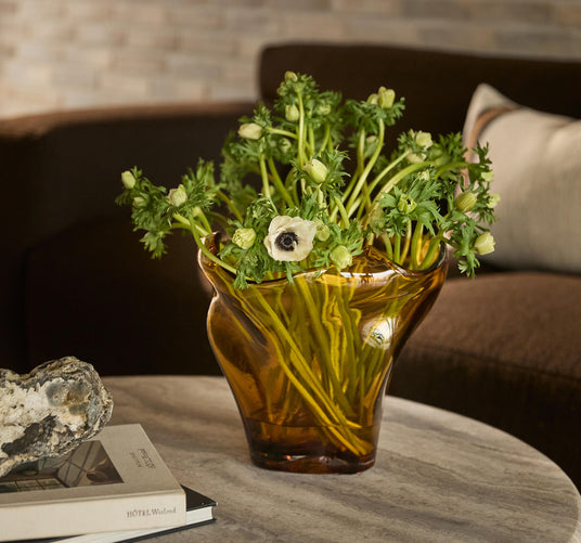 Ravgul glasvase med grønne stilke og hvide anemoner på rundt marmorbord; ved siden af ligger en sten og to bøger, hvoraf en har titlen HOTEL Weekend.