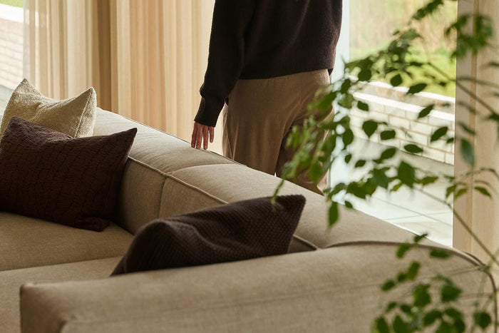 Beige sofa med brune og cremefarvede puder ved lette gardiner; en person står ved vinduet med grønne planter i forgrunden.