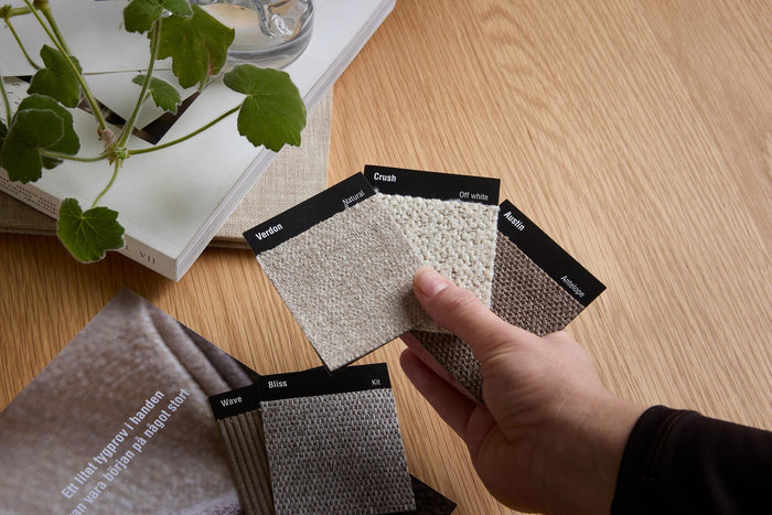 En hånd holder stofprøver med teksten: Verdon – Natural, Crush – Off white, Austin – Antelope; flere prøver ved siden af mærket Wave, Bliss og Kit på et træbord; et magasin ved siden af siger: ”Et lille stofprøve i hånden kan være begyndelsen på noget stort”.
