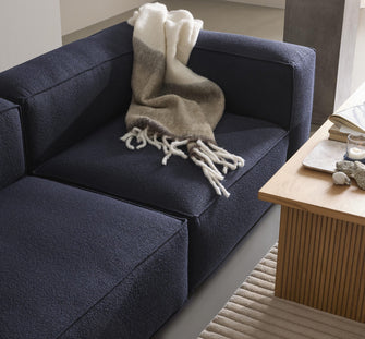 Mørkeblå modulsofa med blød ombré-uldplaid, ved siden af et riflede sofabord i træ med bog og tændt fyrfadslys på bakke, på et beige rillet tæppe i en lys, minimalistisk stue.
