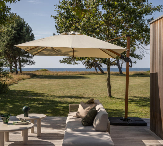 Terrasse med beige loungesofa og små borde under et stort fritstående parasol, udsigt over græsplæne og hav på en solrig dag.