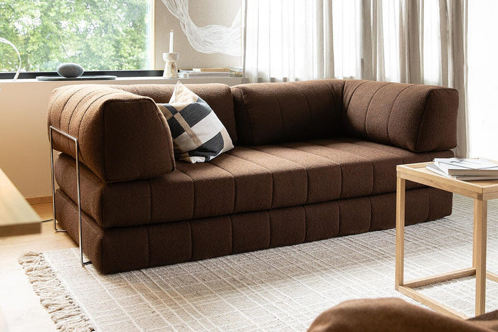Brun modulsofa i lys skandinavisk stue med lyst tæppe, træsidebord, pude og lette gardiner ved vinduet.
