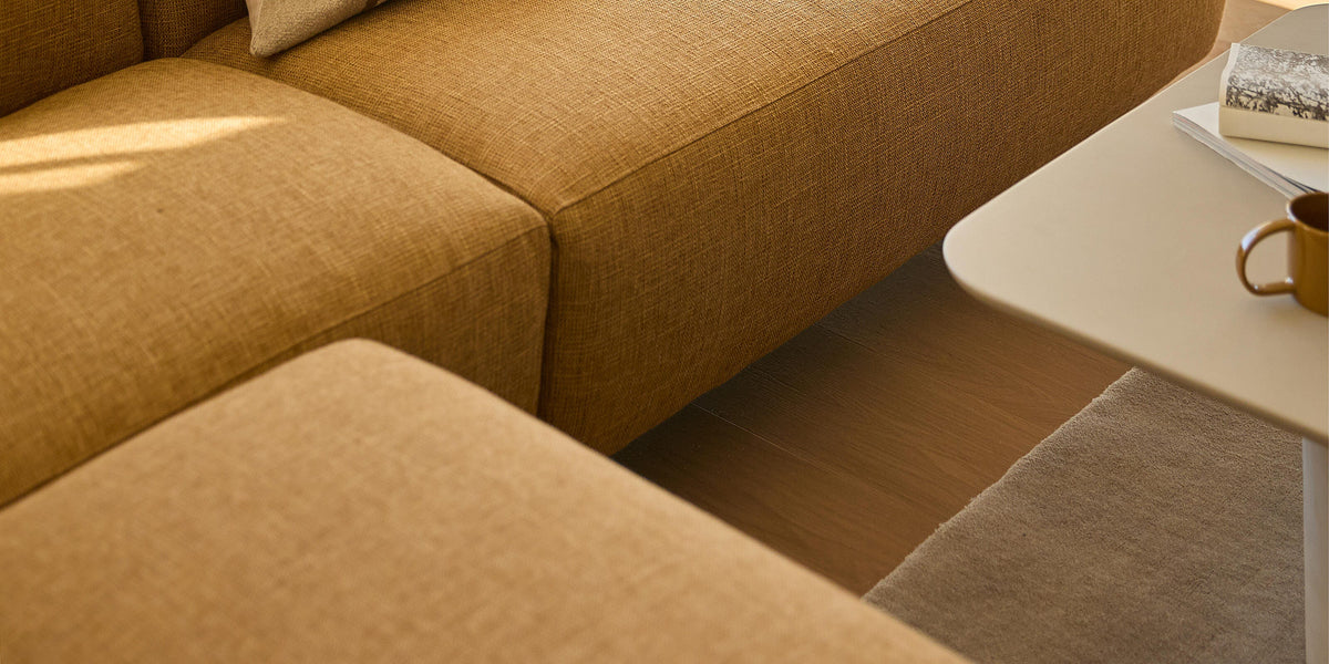 Solrigt stuehjørne med sennepsgul modulsofa og en beige pude med dobbelte striber ved siden af sofabord med kop og bøger.