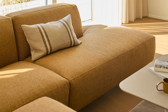 Solrigt stuehjørne med sennepsgul modulsofa og en beige pude med dobbelte striber ved siden af sofabord med kop og bøger.