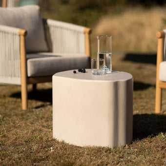 Beige udendørs sidebord i stenlook med karaffel og glas med vand mellem to loungestole på græs i solskin.