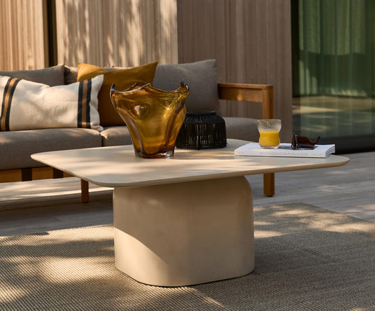Beige udendørs sofabord med skulpturel fod, ravfarvet glasskål, et glas appelsinjuice og en bog med titlen VOL VII; grå sofa med puder i baggrunden på en træterrasse.