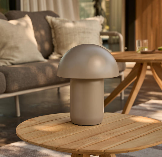 Beige svampeformet bordlampe på rundt træbord på terrasse, sofa med puder i baggrunden.