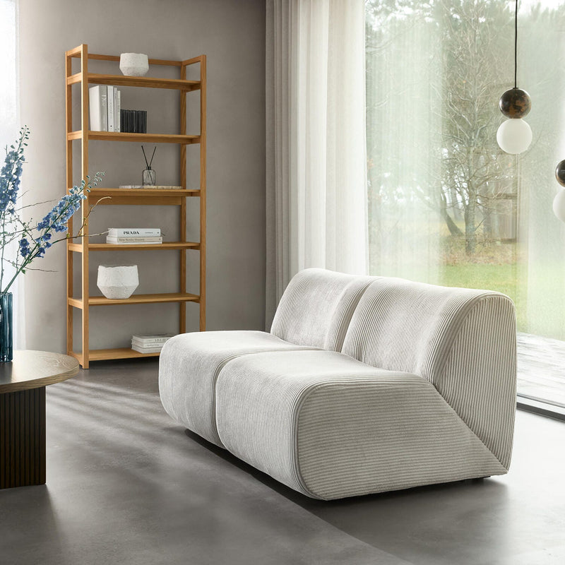 Lys stue med lysgrå modulsofa i fløjl, træreol med bøger inkl. PRADA, blå vase med blomster og runde pendellamper foran store vinduer med lyse gardiner.