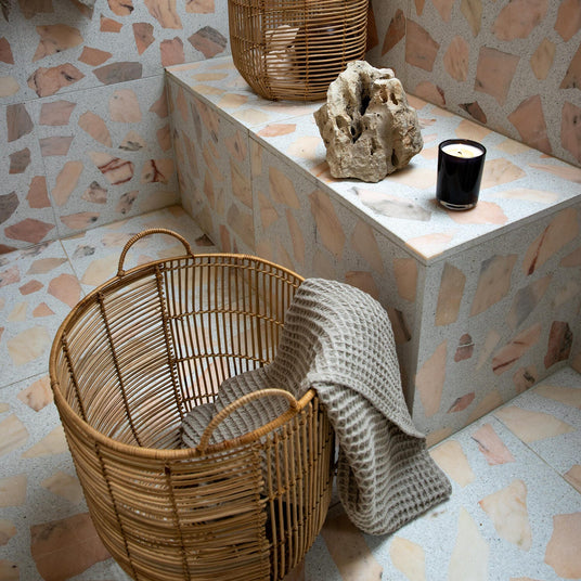 Terrazzo-beklædt badeværelsesniche med to rattan-kurve, beige vaffelhåndklæder, et tændt lys og en dekorativ sten.