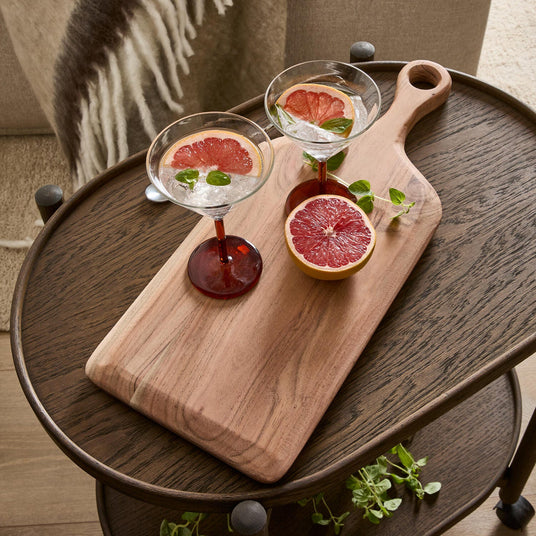 To cocktailglas med skiver af grapefrugt og urter på et skærebræt i træ på et mørkt sidebord, med en halveret grapefrugt ved siden af.
